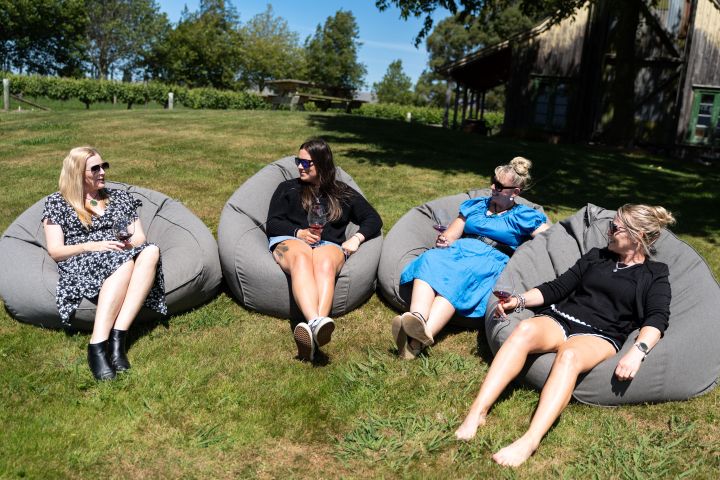 Four women relax on bean bags outdoors, enjoying drinks in a sunny garden with a rustic building in the background.