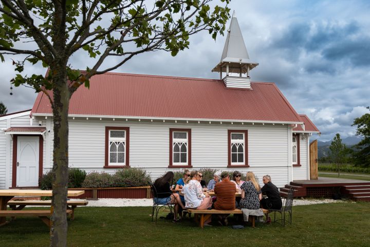 Group of people sitting at picnic tables outside a small white church with a red roof.