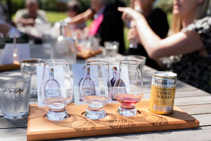 Tray with three glass tumblers and a can of tonic water on a table outdoors with people seated in the background.