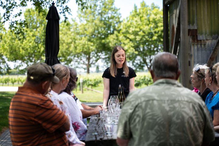 Woman leading a wine tasting outdoors with a group seated at a table.