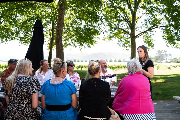 A group of people sitting outdoors under trees, listening to a woman speaking.