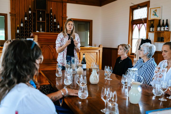 Woman conducting a wine tasting session in a rustic room with seated attendees.